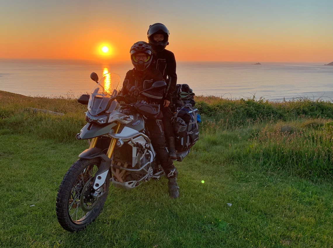 Two women sitting on the Tiger 900 Rally Pro on the coastline around Sennen at sunset