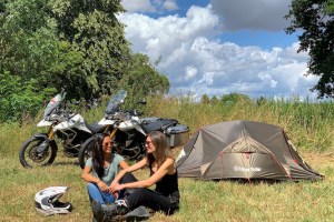 Two motorcyclists sitting in front their ADV Tent in a field and two Tiger 900 Rally Pro motorcycles in the background
